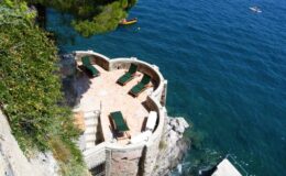 Scenic waterfront terrace with lounge chairs overlooking clear blue water, surrounded by lush greenery and rocky cliffs. Ideal for relaxation and enjoying coastal views.