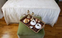 Cozy tea setup on a green ottoman featuring a wicker tray with tea cups, pastries, and decorative candles, set against a softly draped bedspread in a warm wooden room.