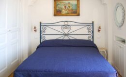 Cozy bedroom featuring a blue bedspread, decorative iron bed frame, and a vintage painting on the wall, with white cabinetry and a round mirror in the background. Ideal for home decor inspiration.
