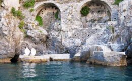 Coastal view featuring rocky cliffs with arched openings and serene turquoise water, showcasing a small stone platform and steps leading into the sea, ideal for swimming or relaxation.