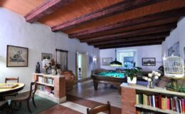 Spacious interior of a rustic living room featuring exposed wooden beams, a billiard table, a cozy reading nook with bookshelves, and a dining area with elegant furnishings. Natural light filters through large windows, enhancing the warm ambiance of the space.