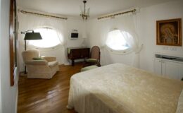 Cozy and elegantly designed hotel room featuring a plush bed, comfortable seating area with armchairs, and stylish decor. Natural light streams through unique round windows, enhancing the inviting atmosphere. Ideal for relaxation and comfort during your stay.