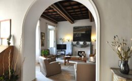 Cozy living room with wooden beams, stylish furniture, and a decorative fireplace, viewed through an archway. Natural light illuminates the space, enhancing its inviting atmosphere.