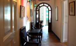 Interior view of a hallway featuring wooden chairs and tables, framed artwork on the walls, and an arched entrance that leads to a brightly lit area, showcasing a welcoming atmosphere.
