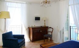 Cozy hotel room featuring a blue armchair, wooden dresser, and a small TV, with large windows adorned with sheer curtains that offer natural light and a view of the outdoors.