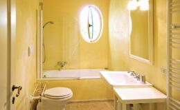 Bright and modern bathroom featuring a yellow color scheme, a bathtub with a shower, a white sink, and a toilet, complemented by natural light from an oval window.