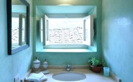 Bright and airy bathroom featuring a large window with a view of rooftops, light green walls, a round sink, decorative plants, and a modern mirror.