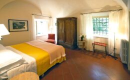 Cozy bedroom featuring a queen-sized bed with yellow accents, wooden wardrobe, and natural light from large windows, creating a warm and inviting atmosphere.