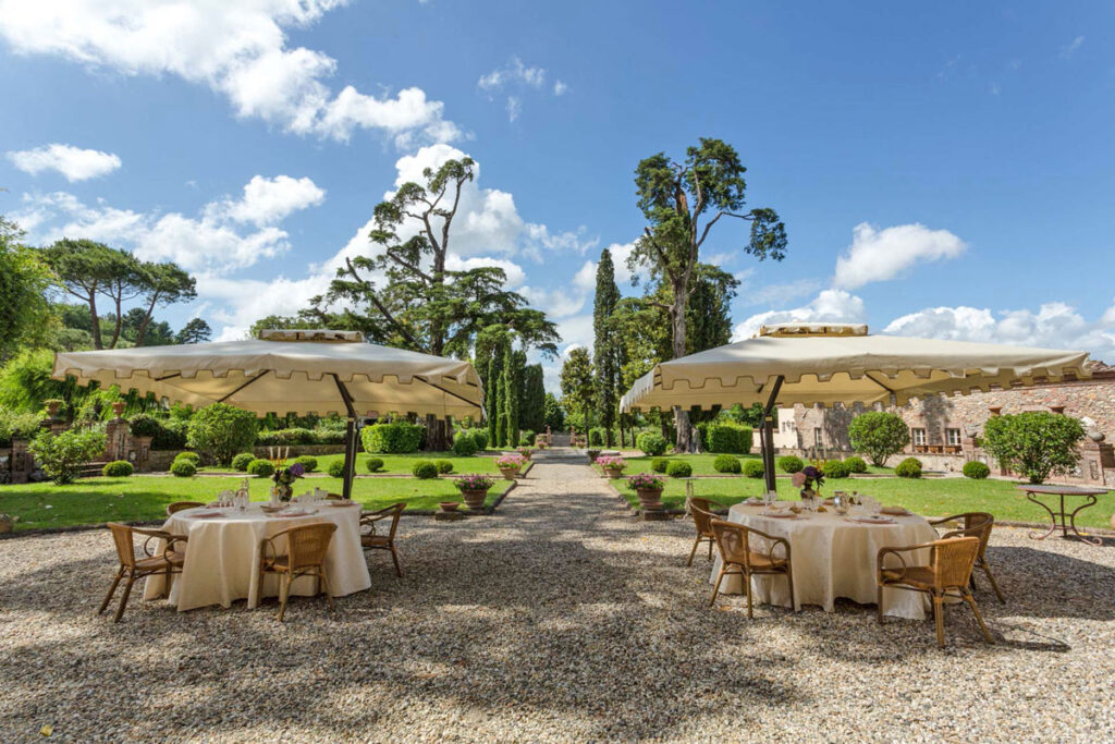 Outdoor dining area in a lush garden featuring two tables set under large umbrellas, surrounded by greenery and decorative plants, under a bright blue sky. | Only on SopranoVillas