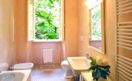 Bright and airy bathroom featuring a large window with greenery outside, a modern sink, toilet, bidet, and elegant decor including a flower arrangement.