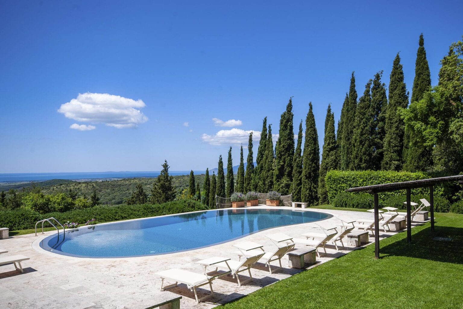 Serene outdoor pool surrounded by lush greenery and cypress trees, with sun loungers and a clear blue sky, perfect for a relaxing getaway. | Only on SopranoVillas