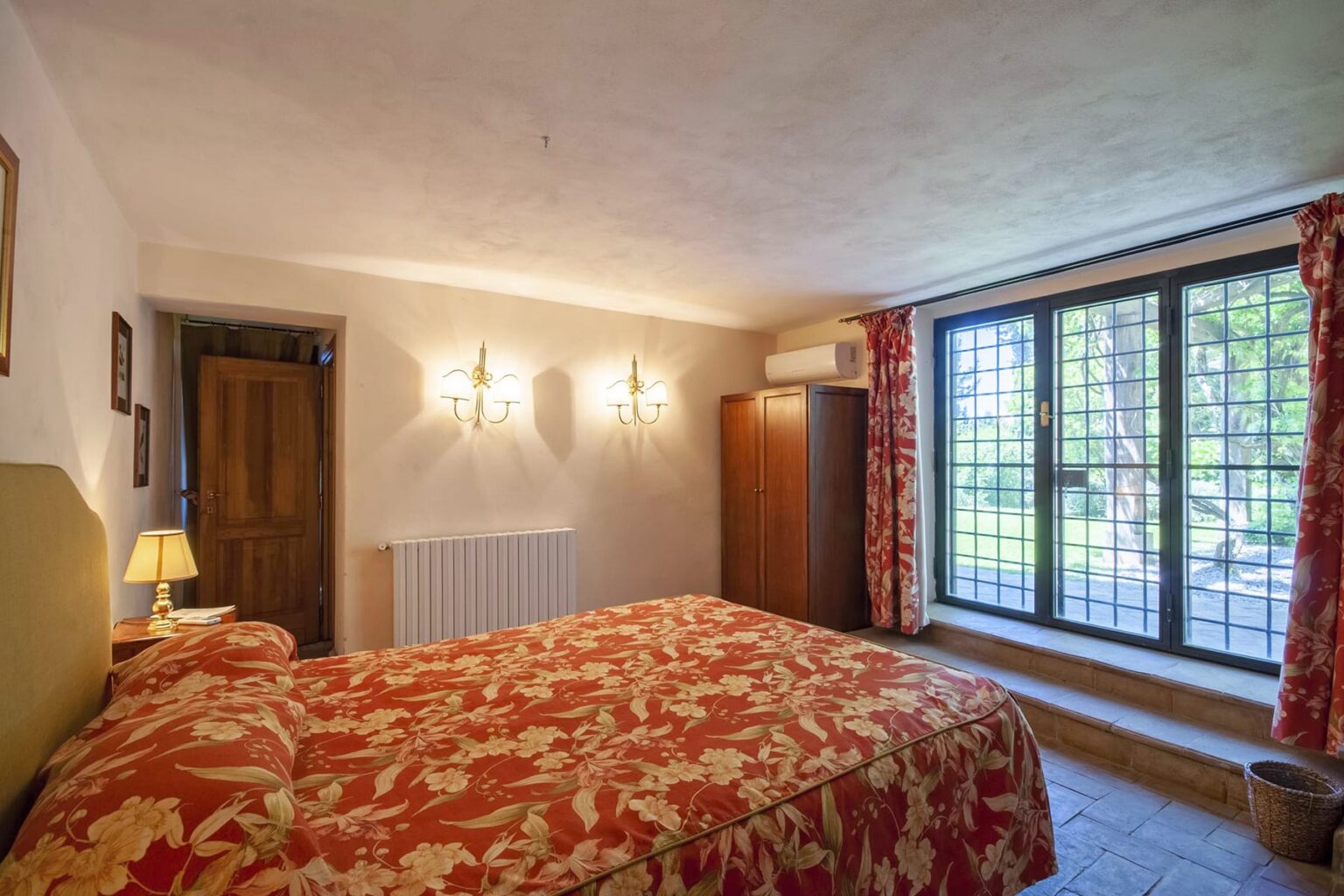 Cozy bedroom featuring a floral bedspread, wooden furniture, and large windows with natural light. The room includes bedside lamps and a wardrobe, creating a warm and inviting atmosphere. | Only on SopranoVillas