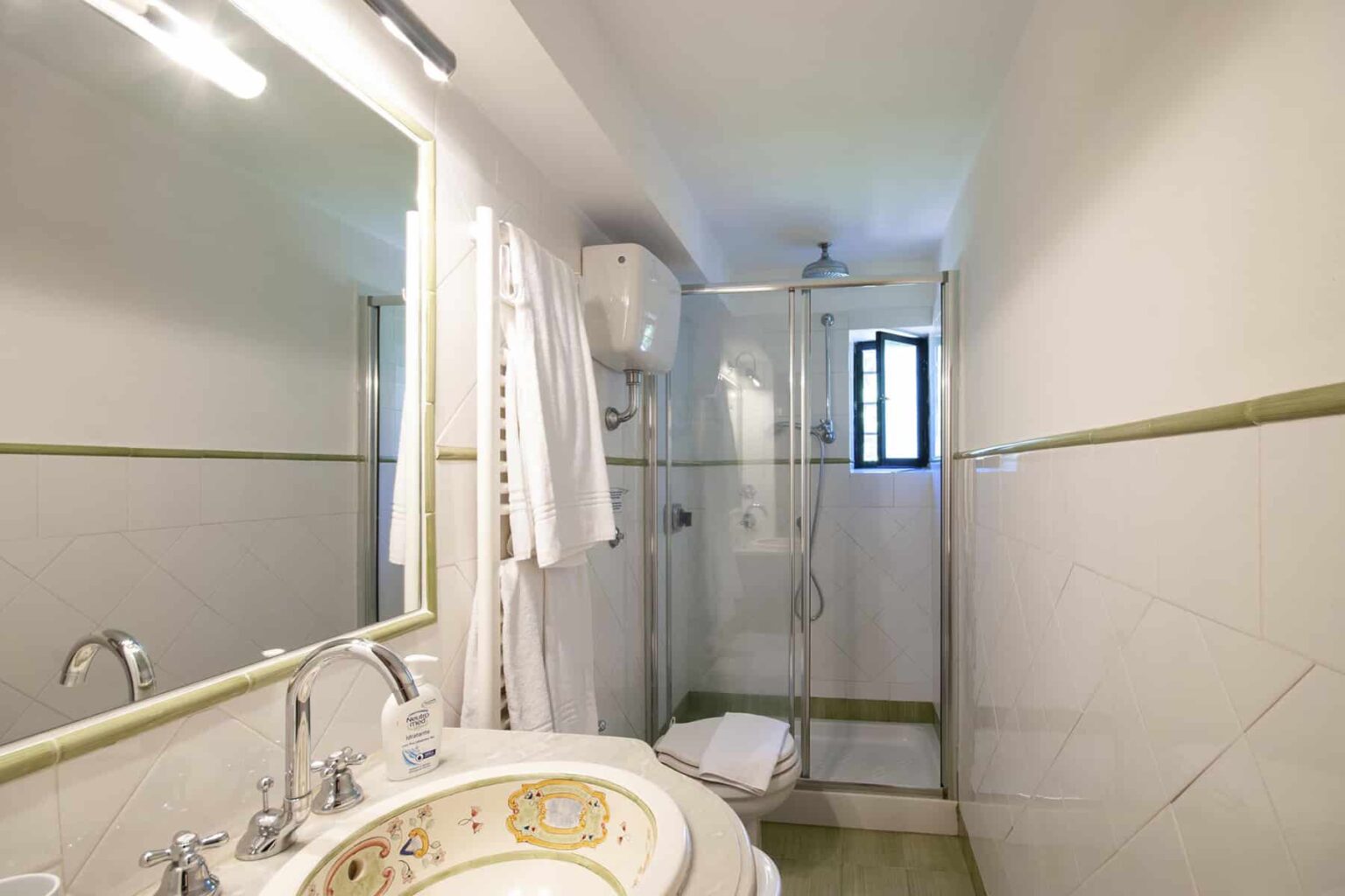 Modern bathroom featuring a glass shower, sink with ornate detailing, and natural light from a window. White and green tile design creates a clean, fresh atmosphere. | Only on SopranoVillas