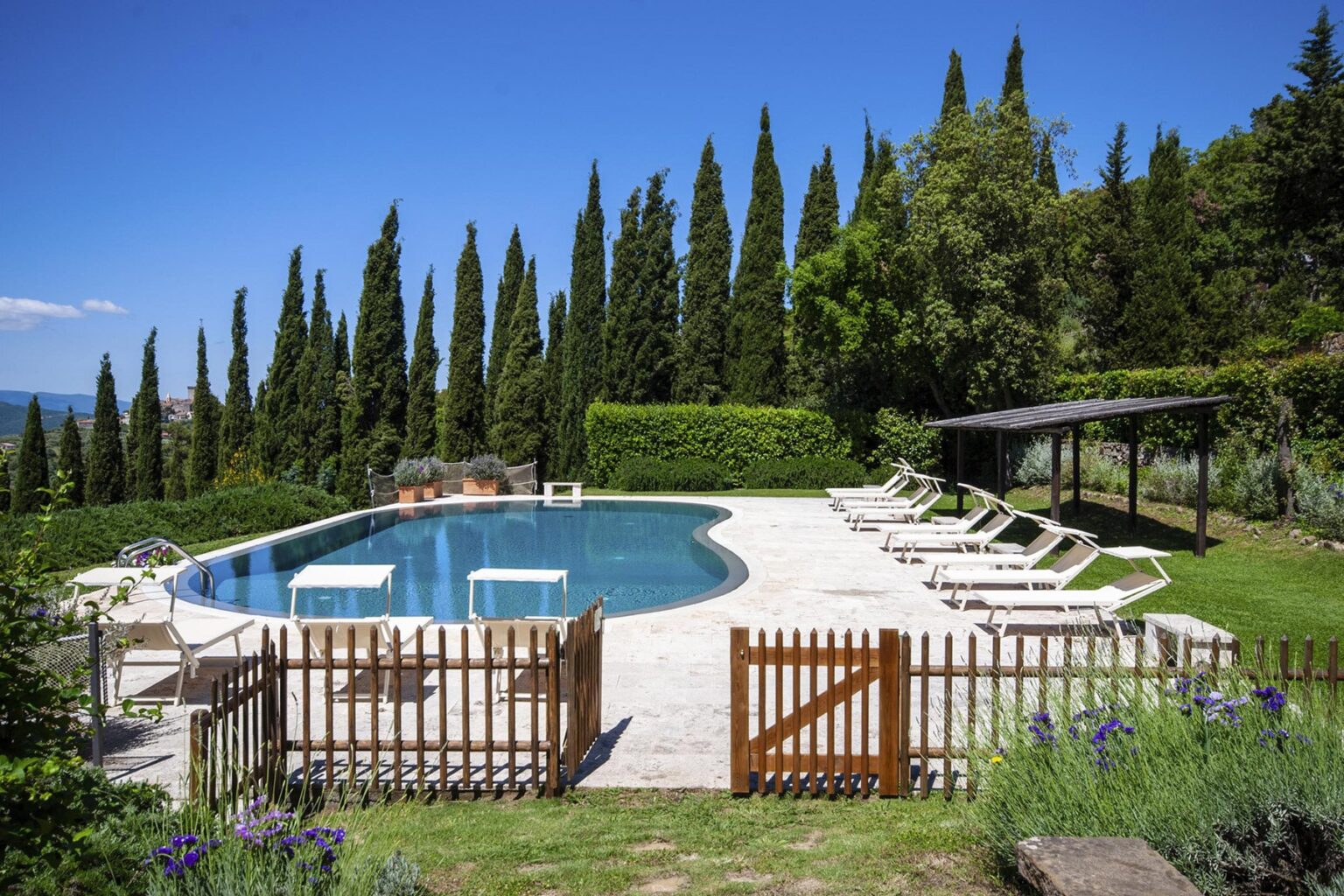 Scenic outdoor swimming pool surrounded by lush greenery and tall cypress trees, featuring sun loungers and a shaded pergola, ideal for relaxation in a tranquil setting. | Only on SopranoVillas