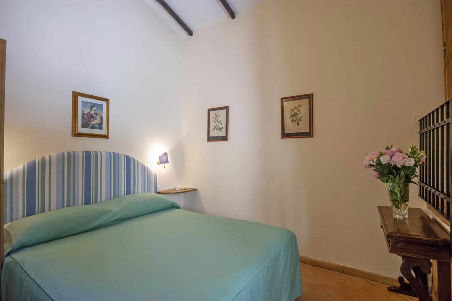 Cozy bedroom featuring a blue striped bedspread, framed artwork on the walls, and a bedside table with a vase of fresh flowers, creating a welcoming and serene atmosphere. | Only on SopranoVillas