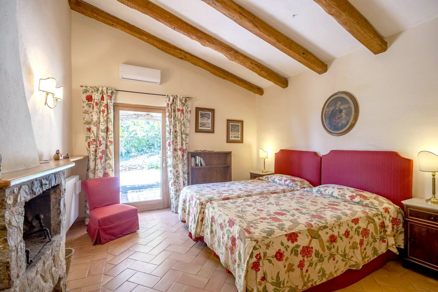 Cozy guest room with two twin beds, floral bedding, wooden beamed ceiling, and a stone fireplace. Features natural light from large windows with curtains and a comfortable seating area. Ideal for a relaxing stay. | Only on SopranoVillas