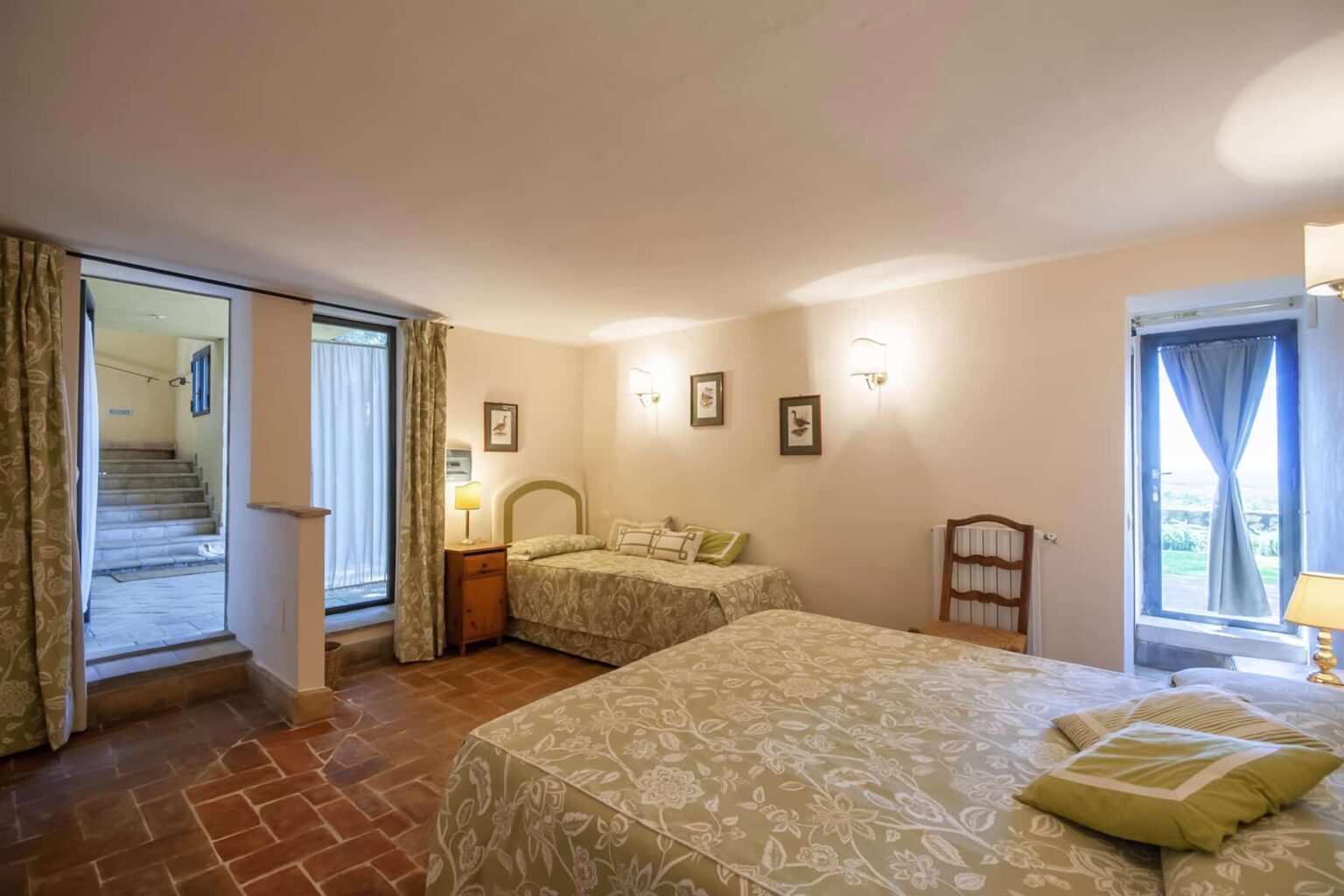 Spacious guest room featuring two beds, elegant decor, and natural light from large windows, with an entrance to a staircase leading outside. | Only on SopranoVillas
