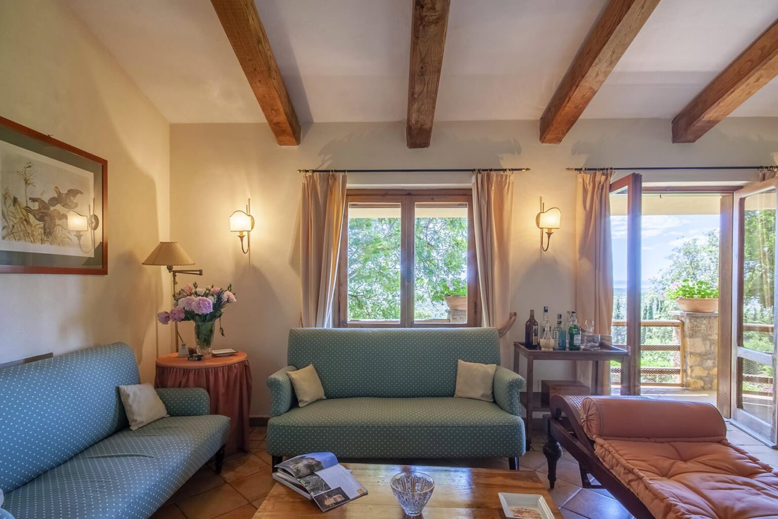 Cozy living room featuring wooden beam ceilings, a green sofa, and large windows with scenic views. The room is decorated with soft lighting, a side table with drinks, and a floral arrangement, creating a warm and inviting atmosphere. | Only on SopranoVillas