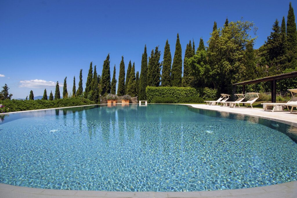 Serene outdoor swimming pool surrounded by lush greenery and tall cypress trees under a clear blue sky, perfect for relaxation and leisure in a tranquil setting. | Only on SopranoVillas