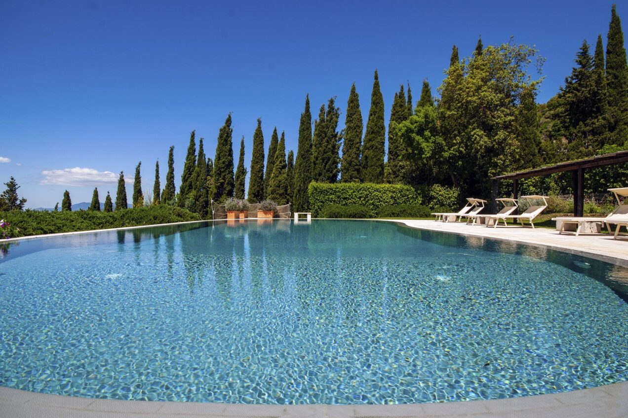 Serene outdoor swimming pool surrounded by lush greenery and tall cypress trees under a clear blue sky, perfect for relaxation and leisure in a tranquil setting. | Only on SopranoVillas