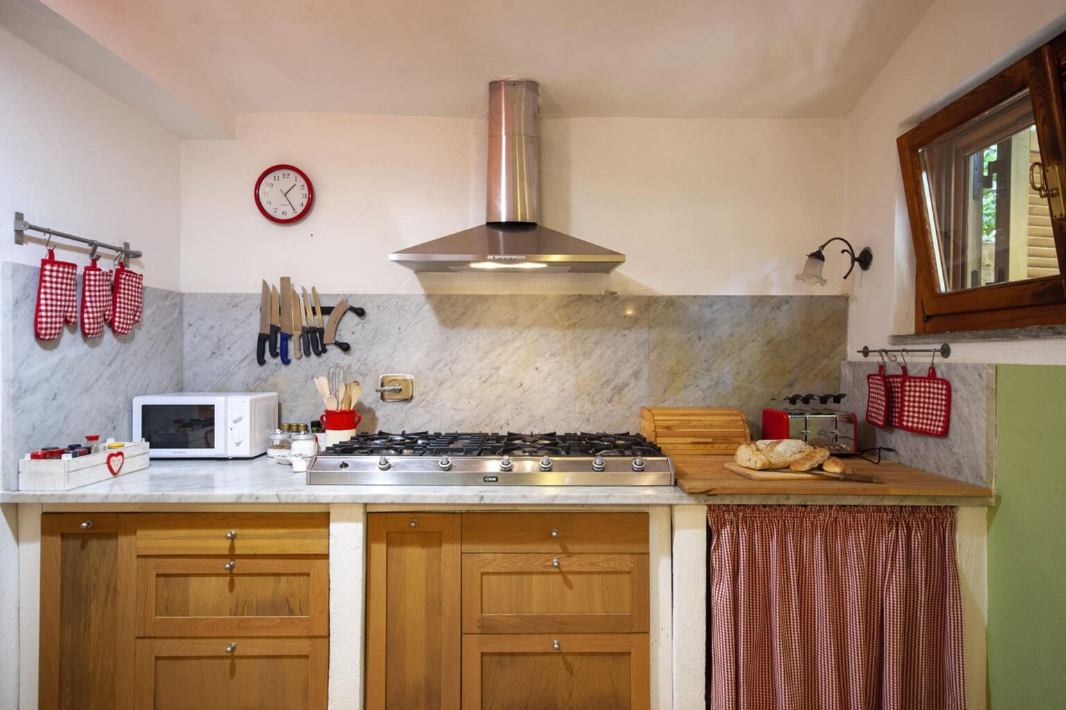 Modern kitchen interior featuring wooden cabinets, a stainless steel range hood, and a marble backsplash. The space includes a microwave, kitchen utensils, and decorative items, with a window allowing natural light. | Only on SopranoVillas