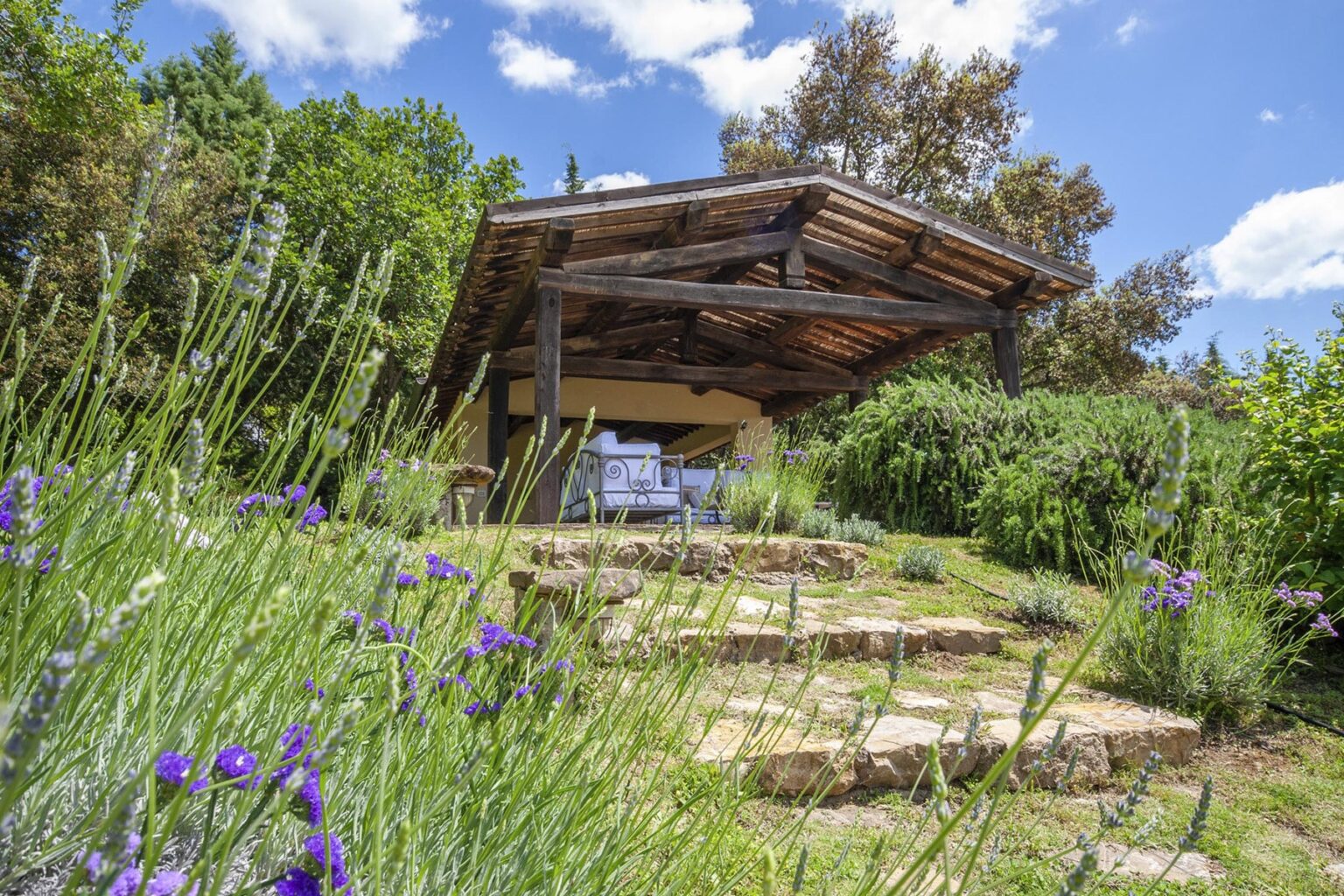 A serene garden scene featuring a stone pathway leading to a wooden gazebo surrounded by lush greenery and blooming lavender flowers under a clear blue sky. | Only on SopranoVillas