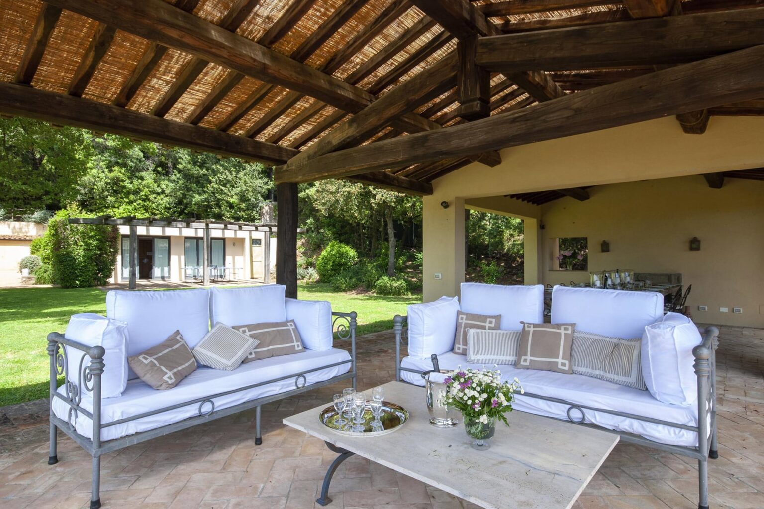 Outdoor living space featuring white cushioned sofas, a coffee table with a floral centerpiece, and a rustic roof, surrounded by greenery and a view of a nearby house. Ideal for relaxation and entertaining. | Only on SopranoVillas