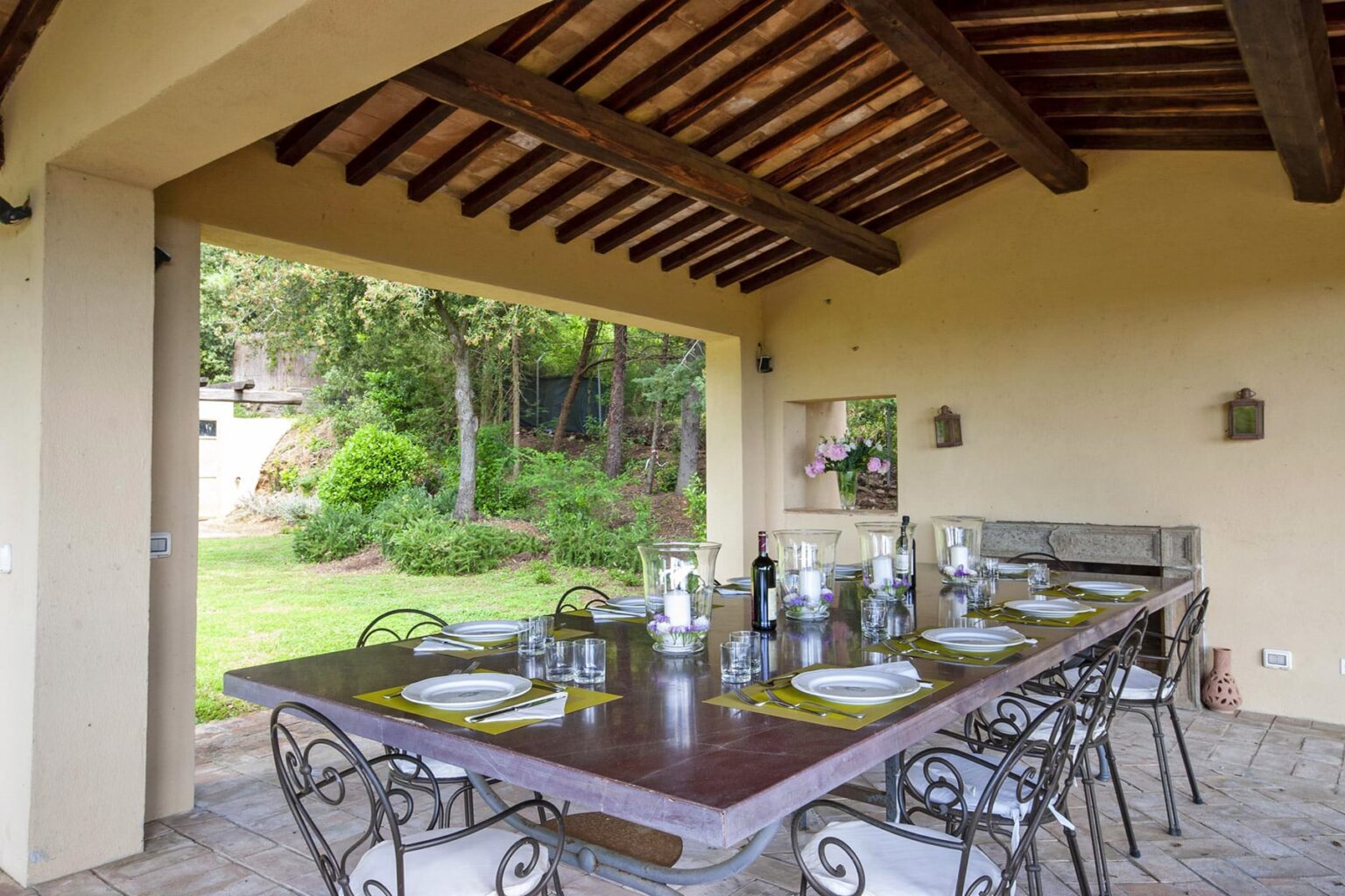 Outdoor dining area with a wooden table set for a meal, featuring elegant tableware and surrounded by lush greenery. The space is covered by a rustic roof, creating a cozy atmosphere for gatherings. | Only on SopranoVillas