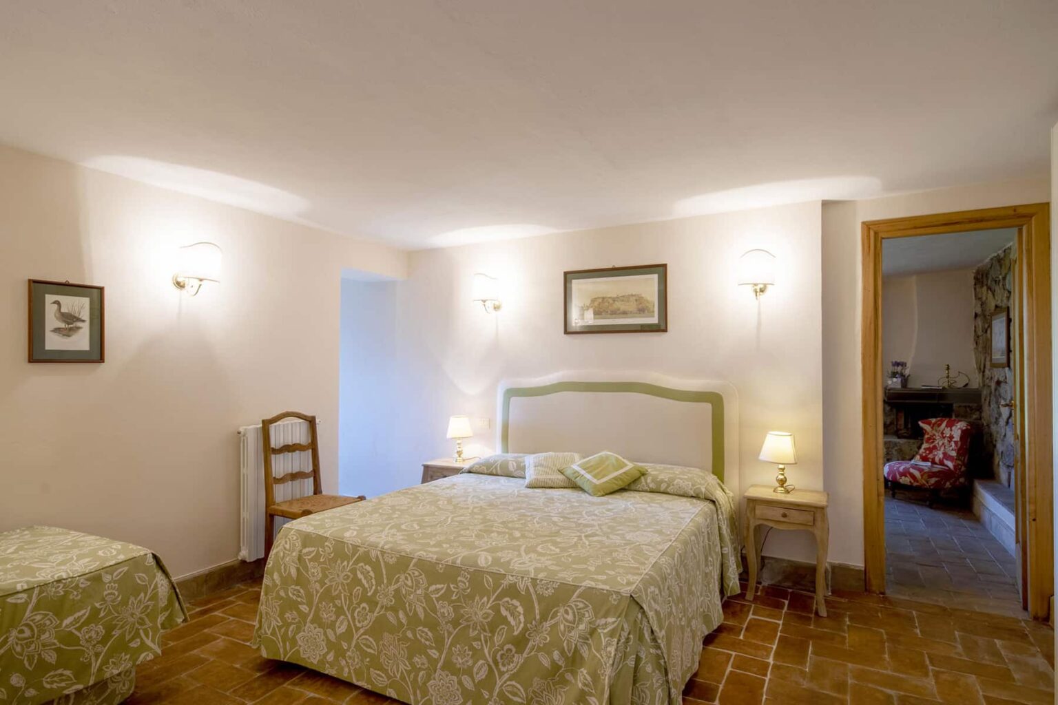 Cozy hotel room featuring a double bed with floral bedding, a bedside table with lamps, and rustic tile flooring. The room includes a vintage chair and an adjoining door leading to a bathroom, creating a warm and inviting atmosphere. | Only on SopranoVillas