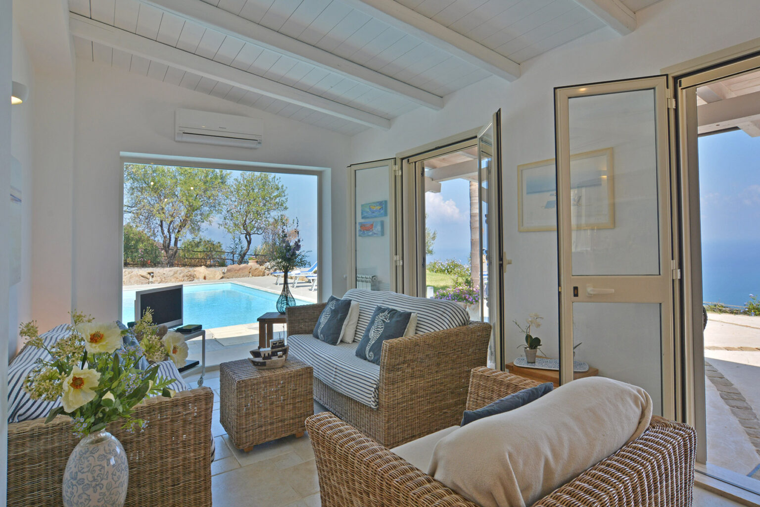 Bright and airy living room featuring wicker furniture, large glass doors opening to a scenic pool view, and coastal decor, ideal for a relaxing vacation setting. | Only on SopranoVillas