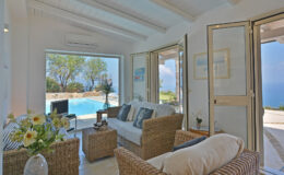 Bright and airy living room featuring wicker furniture, large glass doors opening to a scenic pool view, and coastal decor, ideal for a relaxing vacation setting.