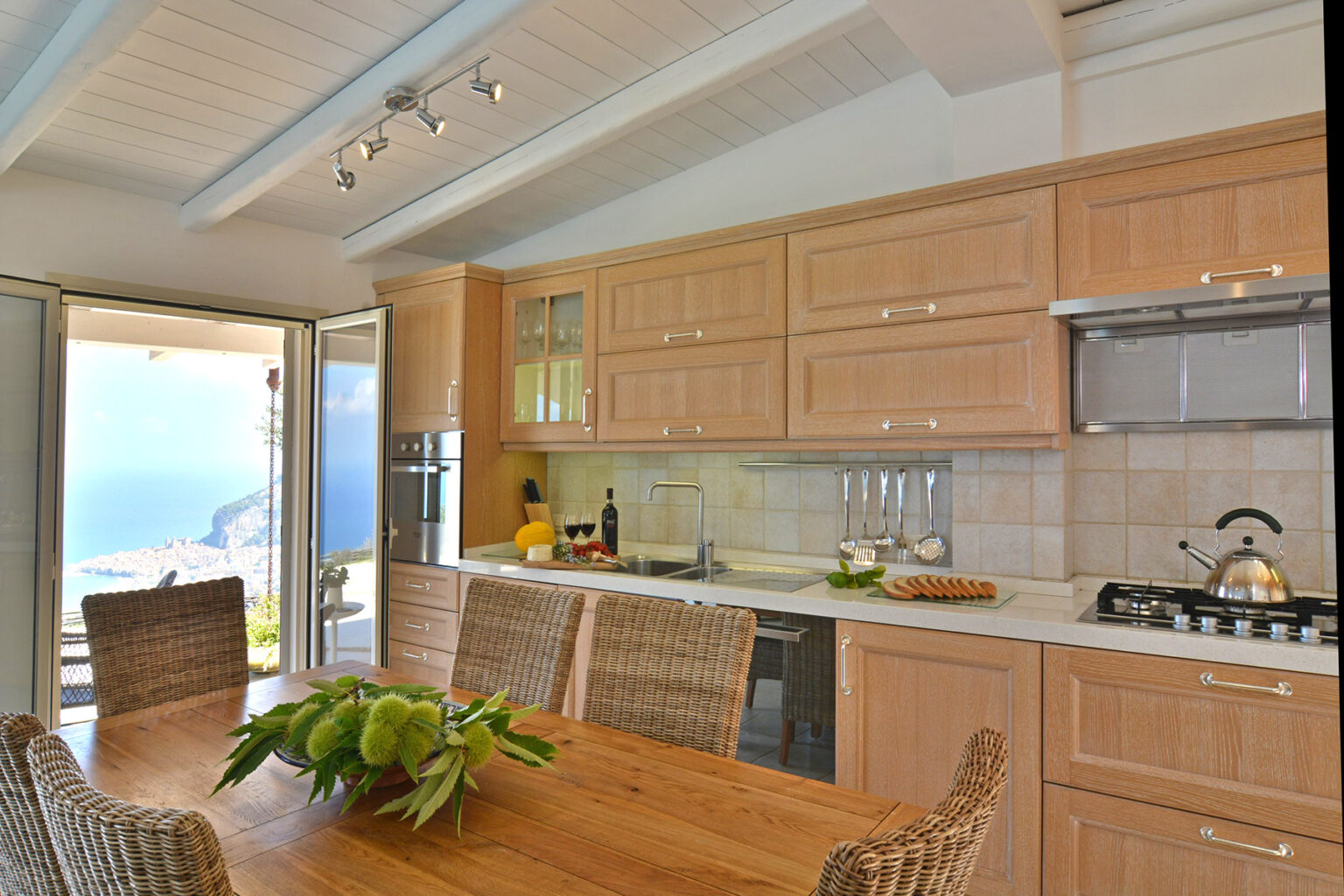 Modern kitchen interior with wooden cabinetry, stainless steel appliances, and a dining table set for six, featuring a scenic view through large glass doors. | Only on SopranoVillas