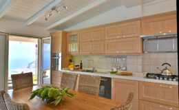 Modern kitchen interior with wooden cabinetry, stainless steel appliances, and a dining table set for six, featuring a scenic view through large glass doors.