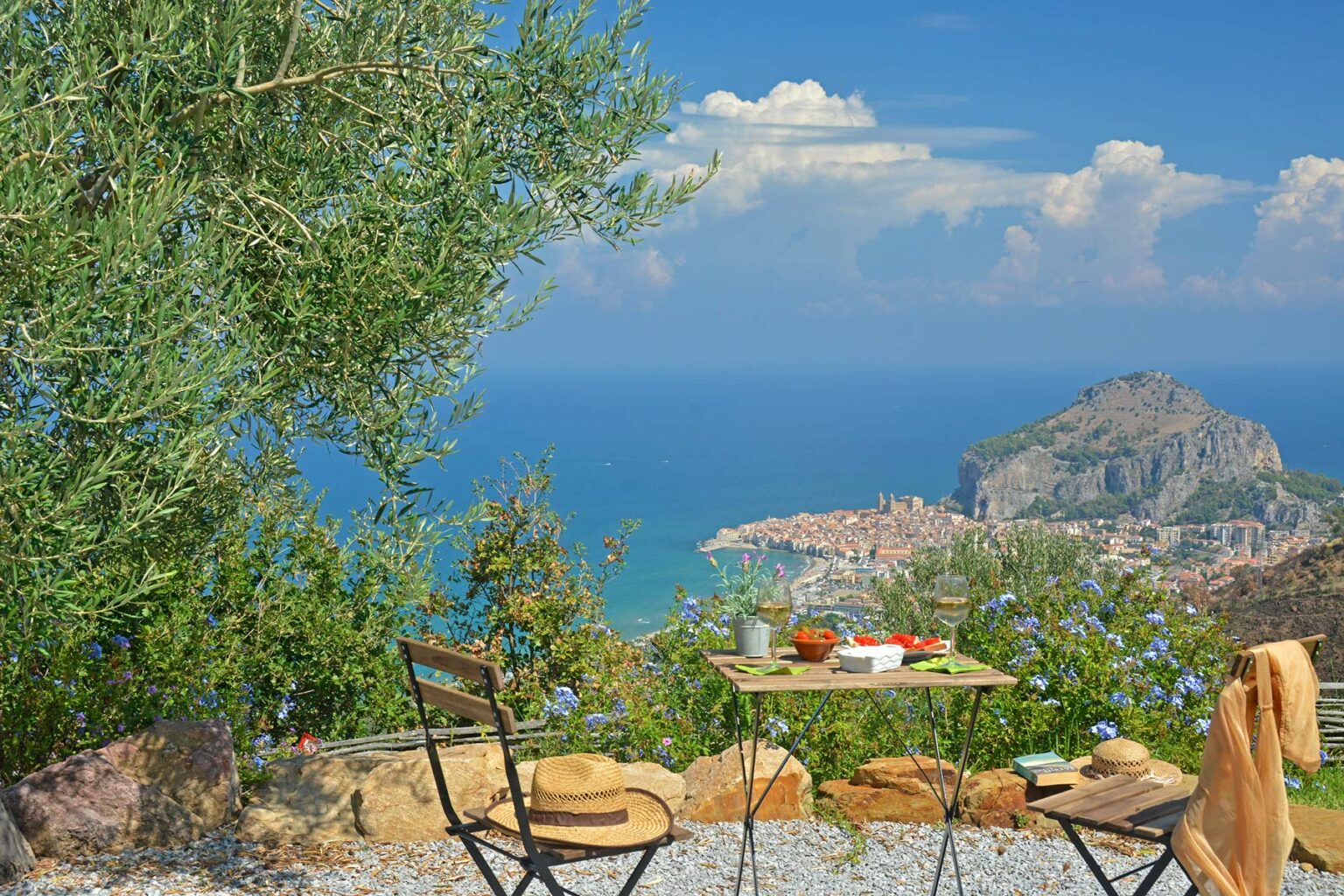 Scenic view from a terrace overlooking a coastal town and the sea, with a table and chairs set among lush greenery and olive trees, ideal for relaxation and outdoor dining. | Only on SopranoVillas