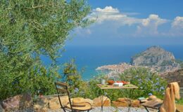 Scenic view from a terrace overlooking a coastal town and the sea, with a table and chairs set among lush greenery and olive trees, ideal for relaxation and outdoor dining.