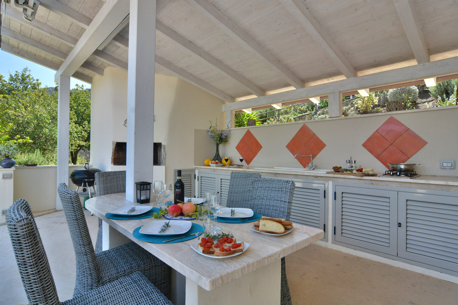Outdoor kitchen with a dining table set for a meal, featuring plates of food, a bottle of wine, and a grill in a serene garden setting. Brightly colored tiles accent the kitchen area, surrounded by lush greenery. | Only on SopranoVillas