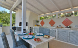 Outdoor kitchen with a dining table set for a meal, featuring plates of food, a bottle of wine, and a grill in a serene garden setting. Brightly colored tiles accent the kitchen area, surrounded by lush greenery.