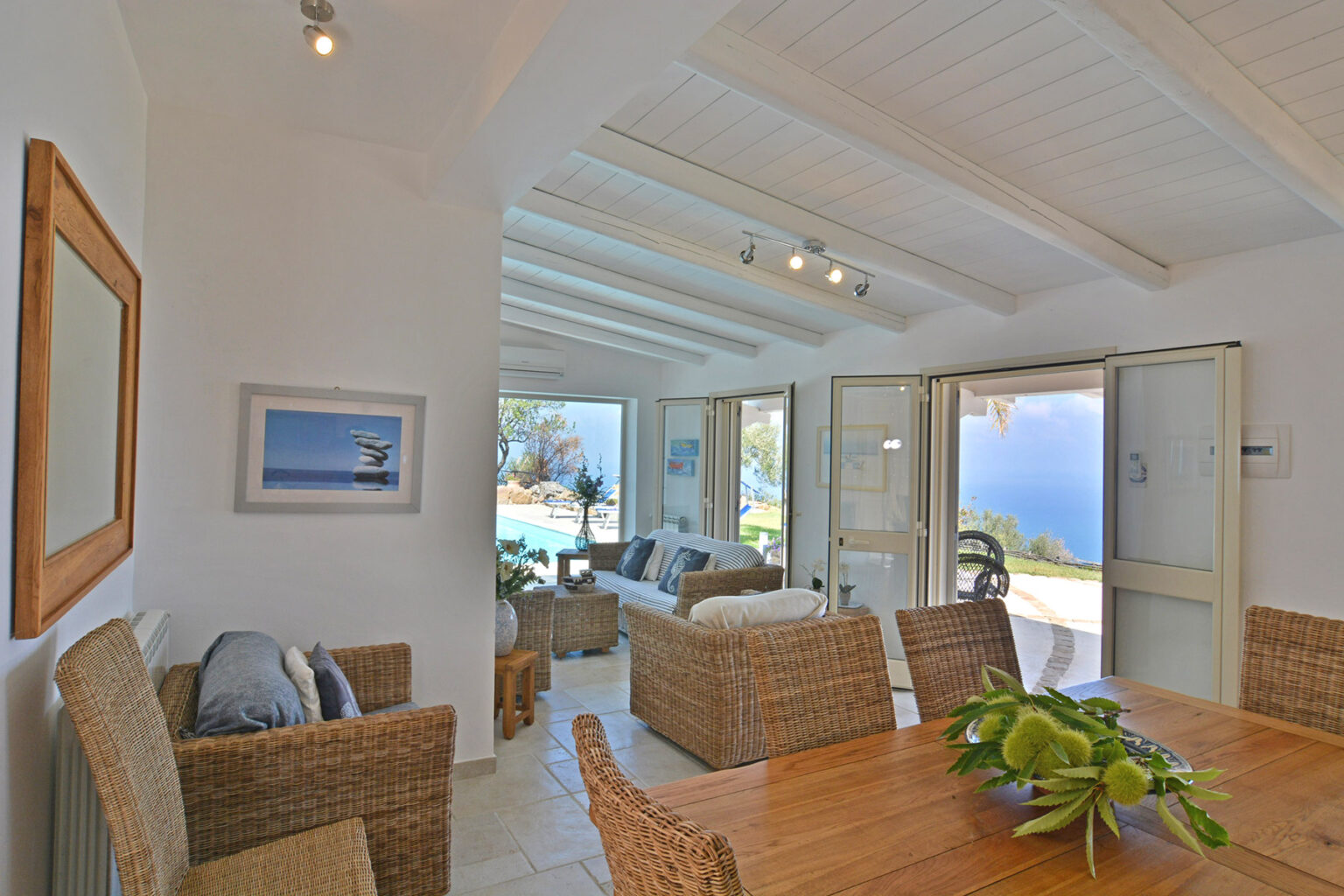 Bright and airy interior of a coastal living space featuring wicker furniture, a wooden dining table, and large windows opening to a scenic view, perfect for a relaxing getaway. | Only on SopranoVillas