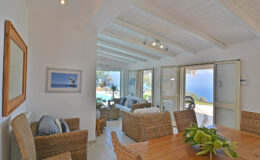 Bright and airy interior of a coastal living space featuring wicker furniture, a wooden dining table, and large windows opening to a scenic view, perfect for a relaxing getaway.