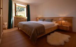 Cozy bedroom featuring a wooden bed with soft bedding, a nightstand with a lamp, and large windows with green curtains, allowing natural light to illuminate the room. A fluffy area rug adds warmth to the wooden floor, creating a serene and inviting atmosphere.