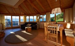 Cozy living room with wooden ceiling, large windows showcasing a scenic view, a comfortable sofa, a dining table with chairs, and warm lighting, perfect for relaxation and family gatherings.