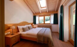 Cozy bedroom featuring a wooden bed with soft bedding, bedside lamp, and large windows with green curtains, providing ample natural light and a serene atmosphere.