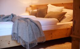 Cozy bedroom featuring a wooden bed with plush pillows and a textured gray throw blanket draped over the side, illuminated by soft ambient lighting. Ideal for showcasing interior design and home decor ideas.