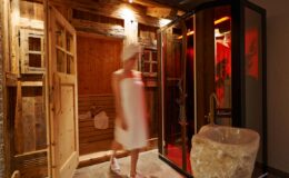 A woman in a white robe walks past a modern shower enclosed in glass, set against a backdrop of rustic wooden walls and a stylish stone bathtub, creating a serene and luxurious bathroom atmosphere.