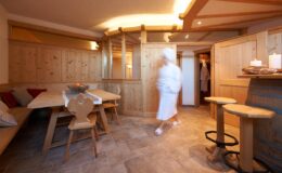 Spa interior featuring wooden walls and furnishings, with a person in a bathrobe walking through a relaxing atmosphere. Soft lighting and a cozy seating area create a serene environment for wellness and relaxation.