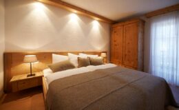 Cozy bedroom featuring a wooden bed with beige bedding, two bedside lamps, and a wooden wardrobe, illuminated by soft wall lighting and natural light from a window.