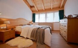 Cozy bedroom featuring wooden beams, a comfortable bed with soft bedding, a nightstand with a lamp, and a wooden dresser, all illuminated by natural light from large windows.