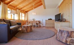 Cozy living room interior featuring a leather sofa, wooden dining table, and natural wood accents. The space is illuminated by large windows and a stylish chandelier, with a round area rug and modern TV unit enhancing the warm atmosphere.