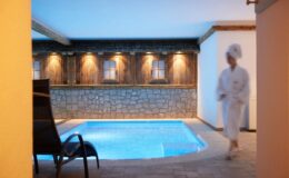 Indoor spa area featuring a tranquil pool, stone walls, and soft lighting, with a person in a robe walking by, creating a serene atmosphere for relaxation and wellness.