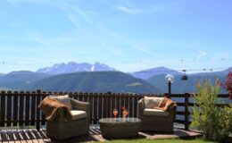 Cozy outdoor seating area with two chairs and a glass table, overlooking a stunning mountain landscape with blue skies and distant peaks. Ideal for relaxation and enjoying scenic views.